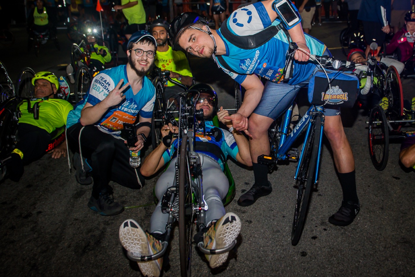 Team Friendship ride-and-roll adaptive cyclists gathered at night with headlamps, volunteers peace-signing — kinetic, inclusive, alive.