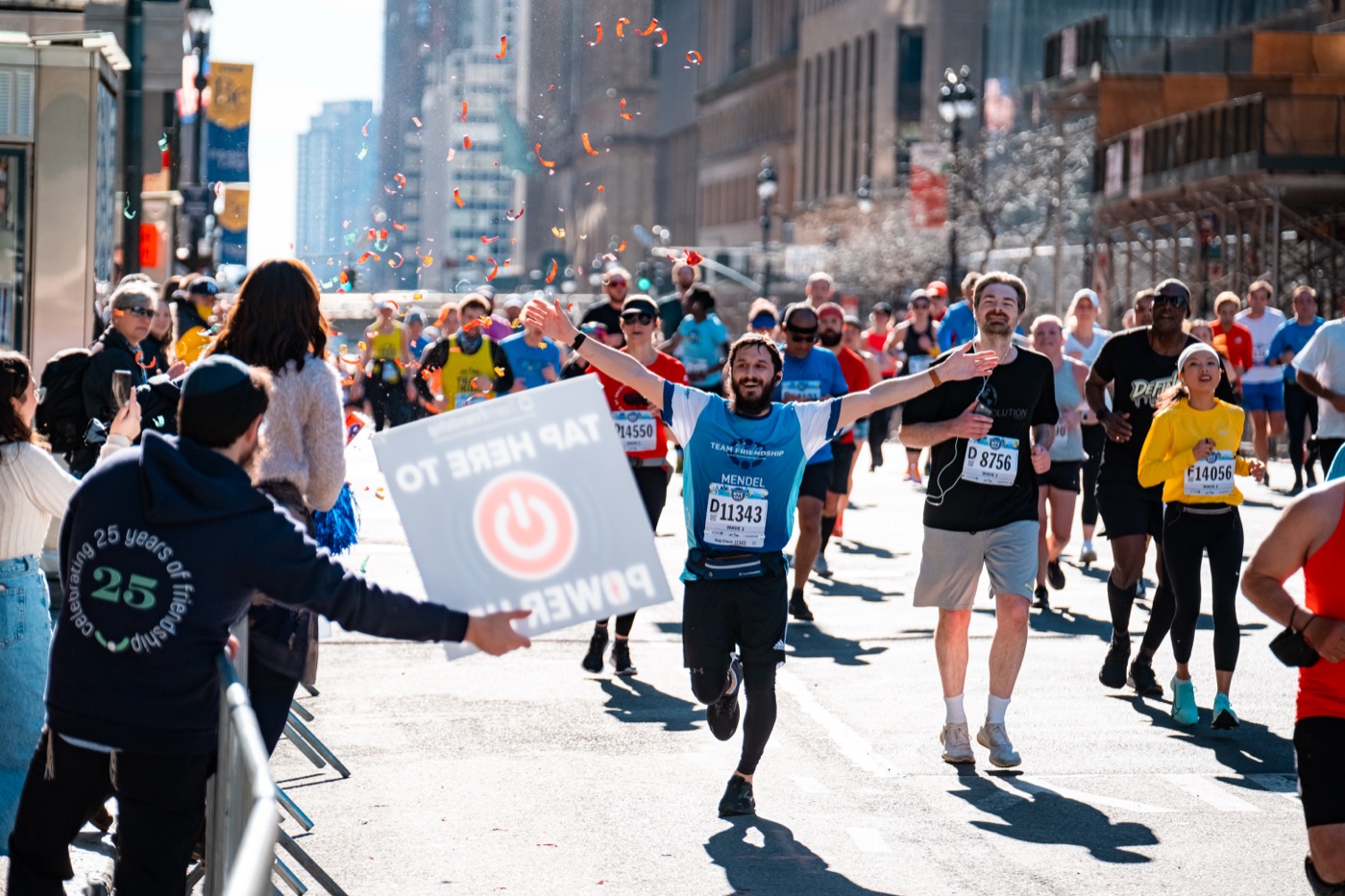 Team Friendship runner crossing the finish with arms raised as confetti falls — joy, arrival, community cheering from the sidelines.