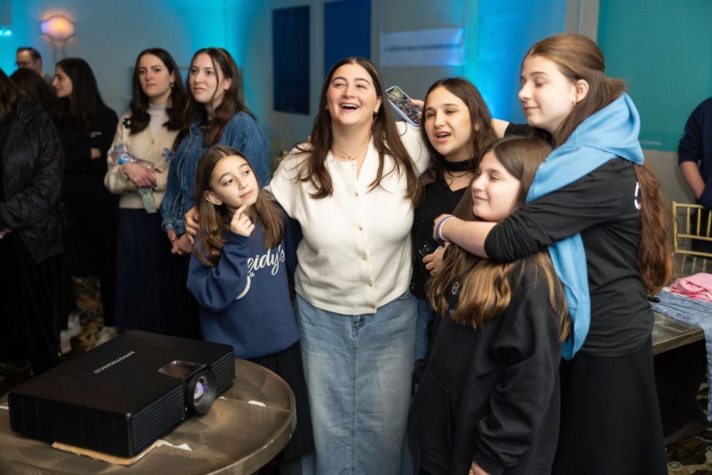 A group of teen girls laughing together at an indoor Friendship Circle event, warm light, arms around each other — community.