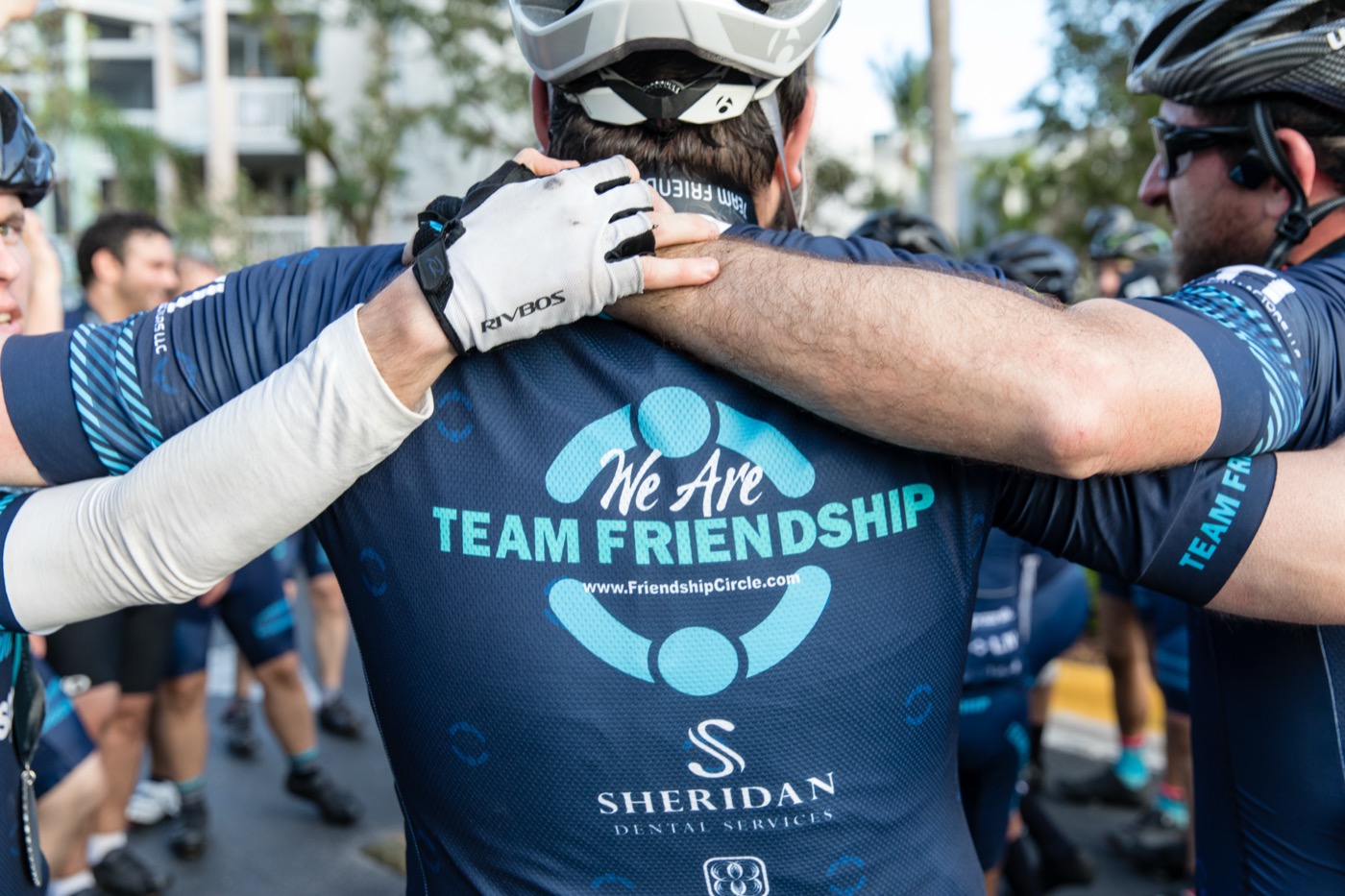 Close crop of Team Friendship cyclists embracing before the ride — jerseys visible, hands on shoulders — camaraderie, unposed.