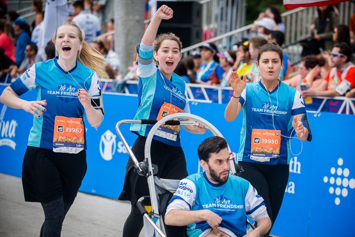 Team Friendship runners in matching kit pushing an adaptive chair at a race, fists up, cheering — the team moment.
