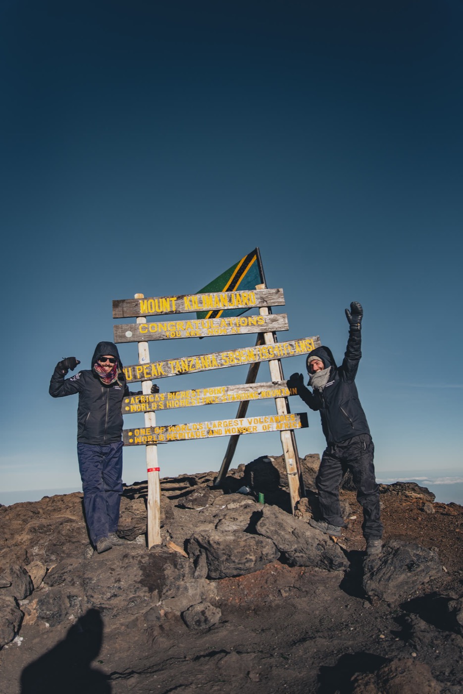 Two climbers fists raised at the Kilimanjaro summit sign under a clear blue sky — triumph, shared, earned.