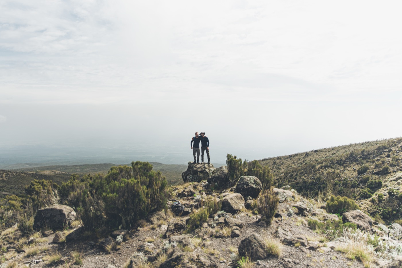 Two hikers standing on a rocky ridge above a vast valley — small against the scale of the landscape, companionable, about to begin.