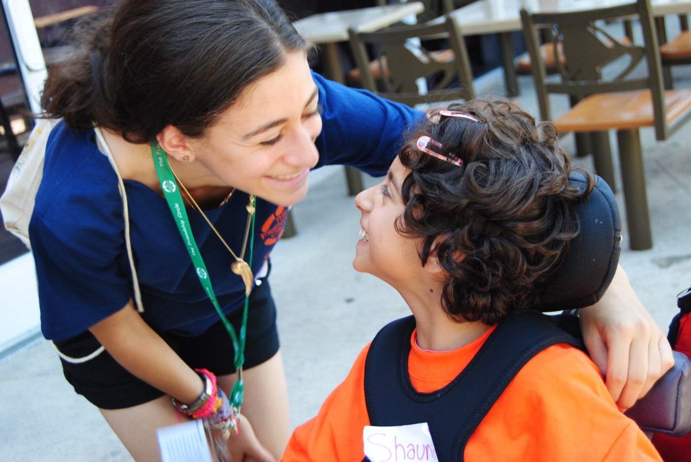 A teen volunteer and a child in a wheelchair share a laughing eye-to-eye moment outdoors — the Friendship Circle core moment. Joy-forward, not pity.